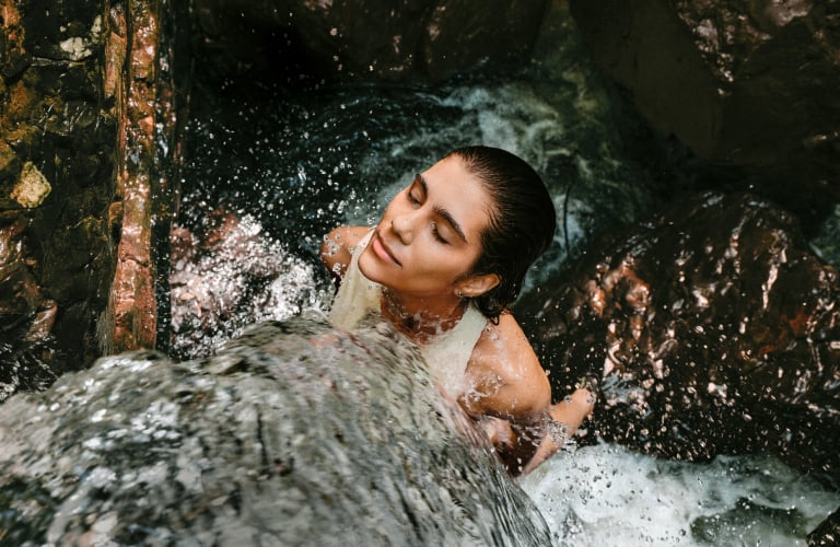Foto de mulher em uma queda-d’água, cercada por água corrente, pedras e vegetação em um ambiente de floresta.
